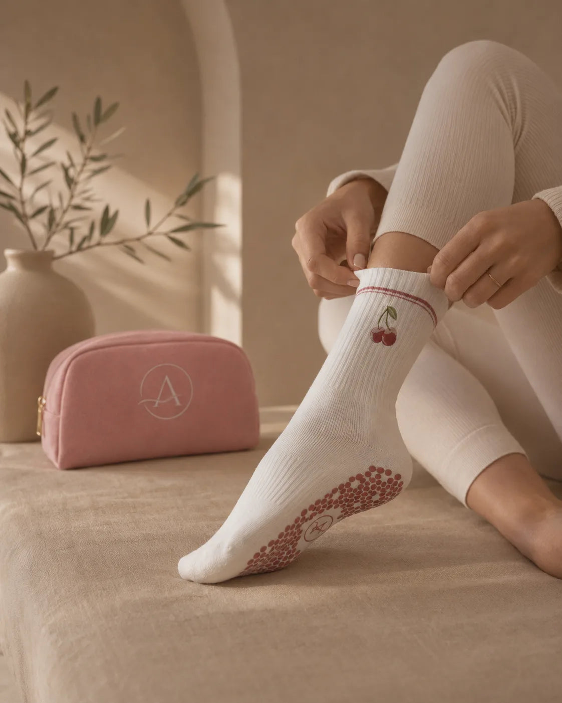 Hands pulling on an Aramé ribbed grip sock with a small embroidered cherry and anti-slip sole, beside the monogrammed dusty-pink studio pouch